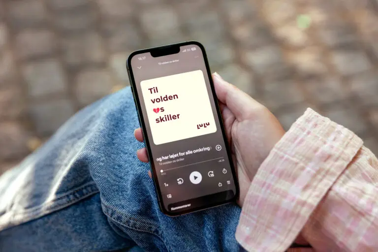 Håndholdt mobiltelefon med en episode af LULUs podcast “Til volden os skiller” afspillende på skærmen, fotograferet udendørs