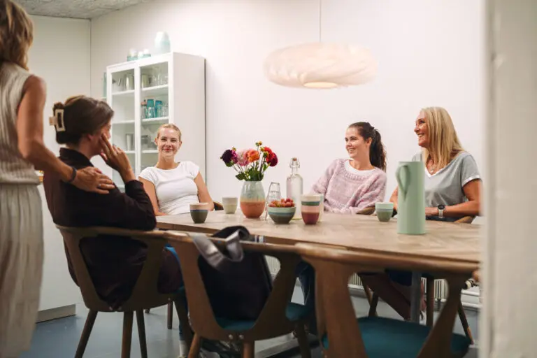 Gruppe af kvinder samlet omkring et bord i hjemlige omgivelser, hvor de smiler og taler sammen