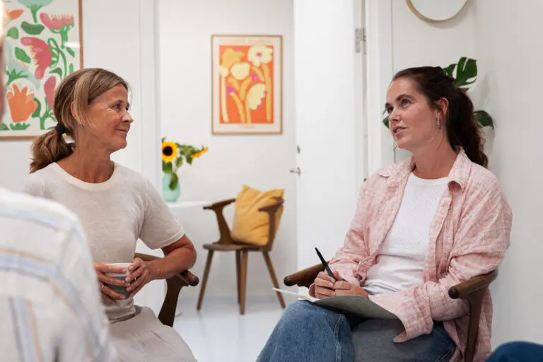 To kvinder i samtale sidder på stole i et lyst lokale. Den ene kvinde har en notesbog og pen, og der ses kunst og møbler i hjemlige omgivelser.